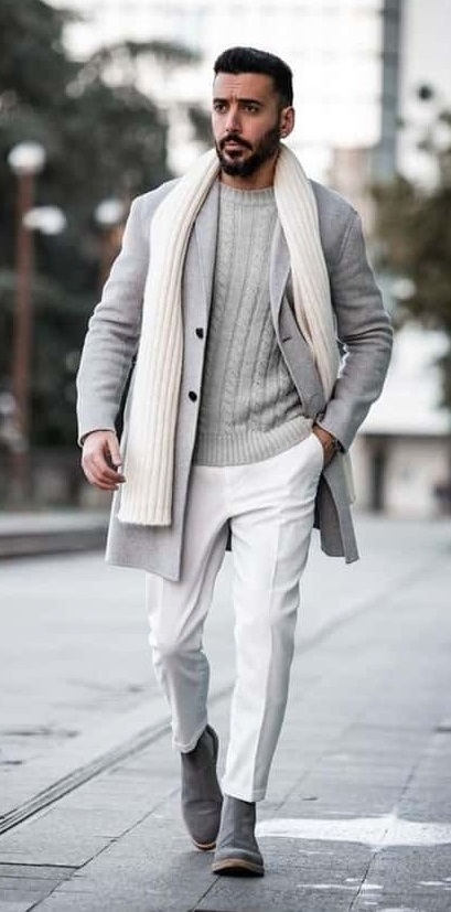 A grey outfit that has grey colored long overcoat and cream scarf. Also wearing a grey sweater inside and grey boots