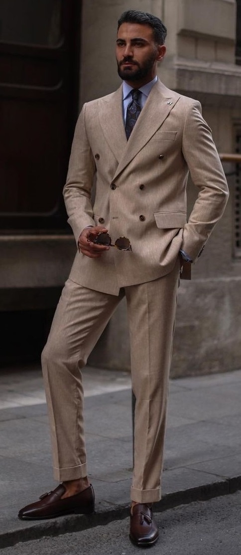 double breasted beige suit with blue shirt and a printed tie, holding shades in hand_