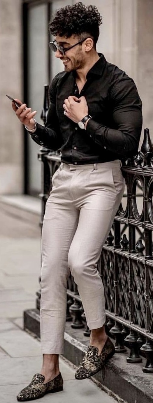 black shirt accessorized with suedes, watch and shades, happily standing with a mobile